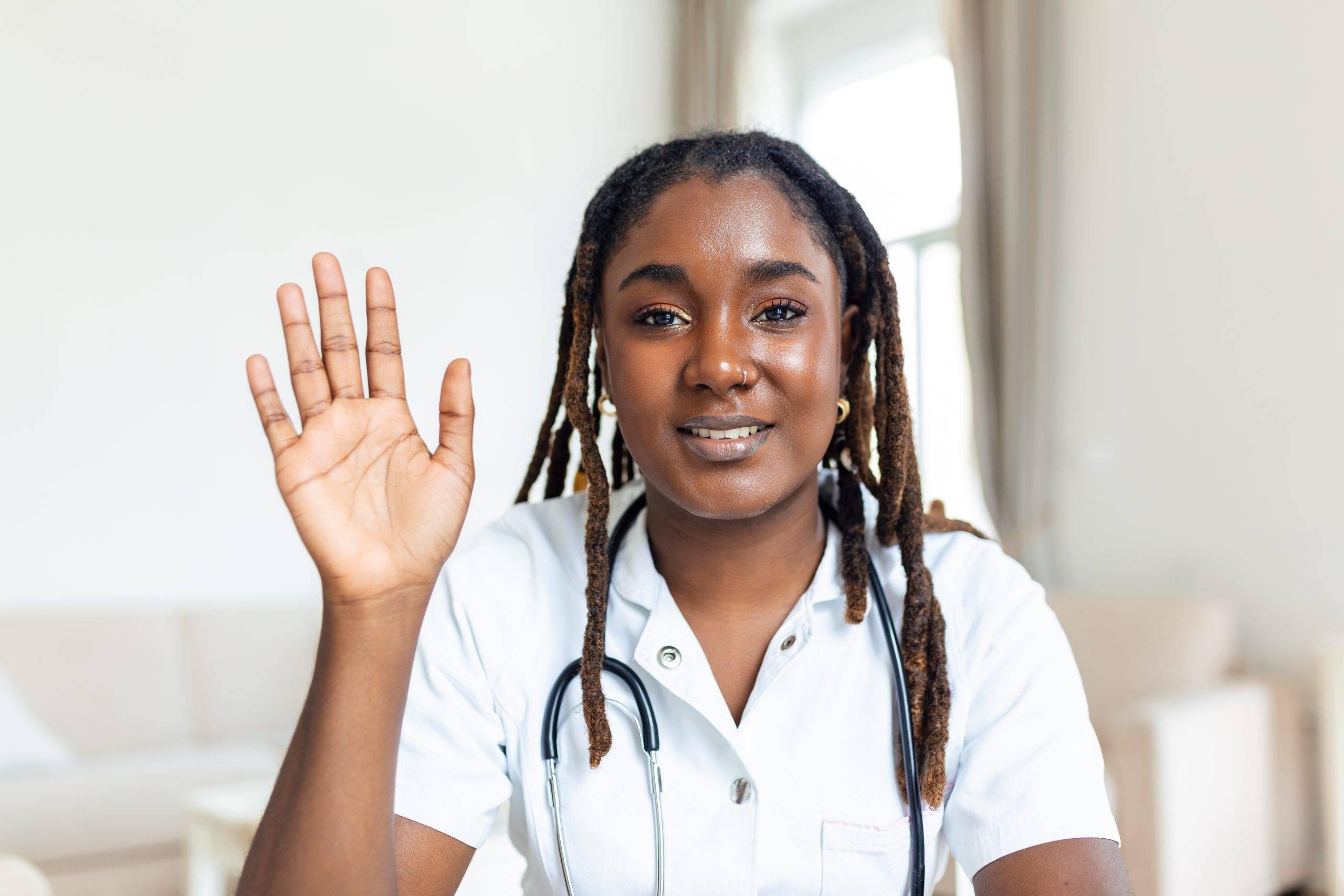 African female doctor talking online with patient, making video call, looking at camera, young female wearing white uniform with stethoscope speaking, consulting and therapy concept