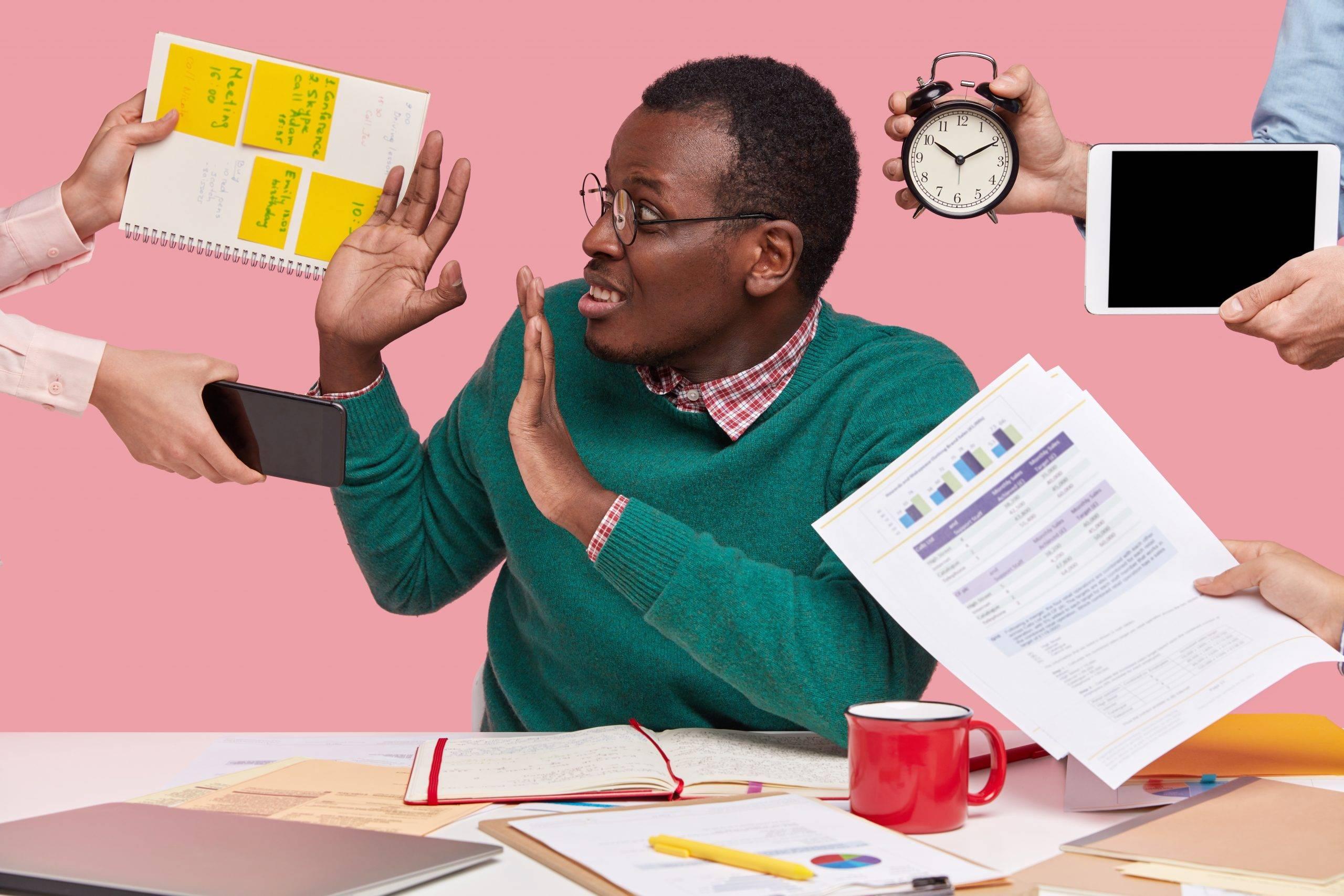 Stressed dark skinned man has much work in office, asked by many people at one time who remind about preparing business project, hold alarm clock, notepad with records, sits at desktopStressed dark skinned man has much work in office, asked by many people at one time who remind about preparing business project, hold alarm clock, notepad with records, sits at desktop