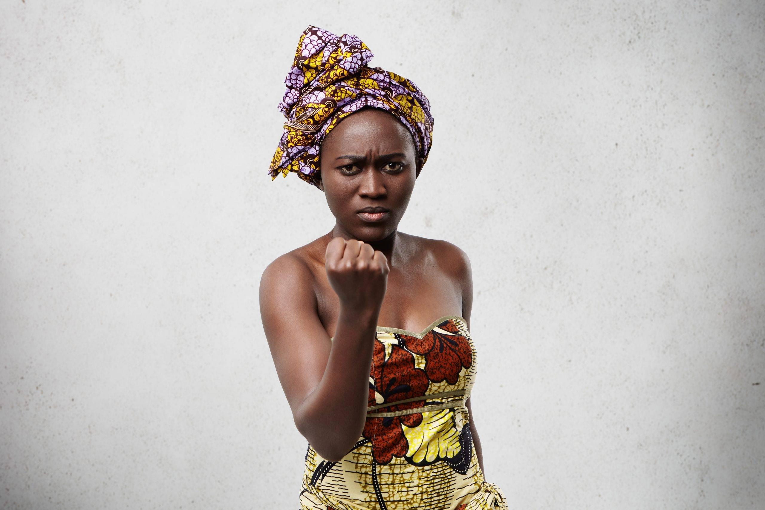 Mental health peoblems in Africa: Menacing black woman with big eyes and full lips wearing colorful scarf on head and dress showing her fist looking angry and furious at camera threatening with gesture. Emotional dark-skinned woman